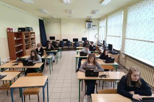 Uczniowie klasy siódmej siedzący w sali informatycznej rozwiązują konkurs przy laptopach.